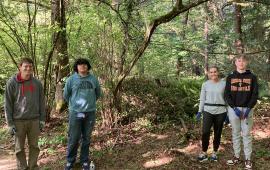 Four volunteers standing in front of a large pile of ivy that they removed, in a forest with dappled light.