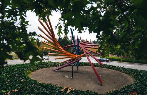 A wing-like public artwork made of colorful steel beams is perched on a round berm in a park on Mercer Island 