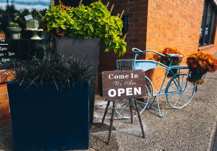 A blue bicycle with flowers in the basket is parked next to a sign that reads "Yes We're Open" outside a flower shop.