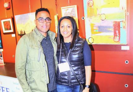 A man and woman smile in front of a wall of paintings at an art gallery reception