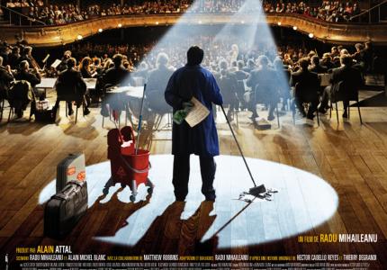 A custodian with a mop and mop bucket stands on a stage looking out a full audience in a big concert hall