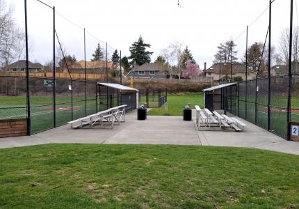 A playfield that includes a cement walkway located between two softball fields and featuring two sets of metal bleachers 