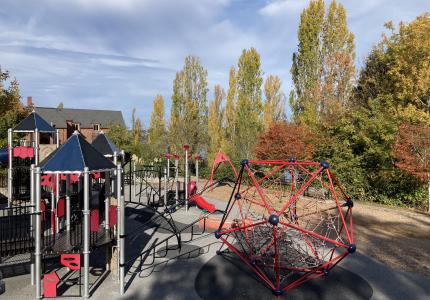 A play area features red, blue, and gray play equipment, including climbing structures, ropes, slides, and a zip line.