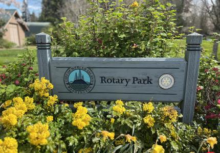 A wood sign reading Rotary Park, surrounded by yellow flowers. 