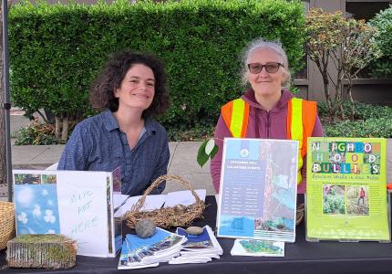 Two people posing at a table of pamphlets about removing non-native weeds.