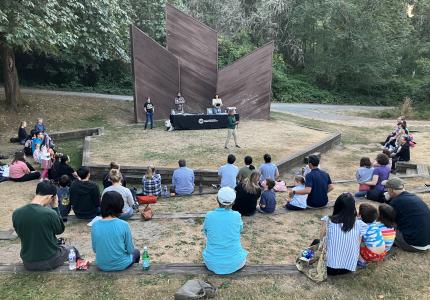 A group of people sitting in front of an amphitheater, with one speaker on stage.  