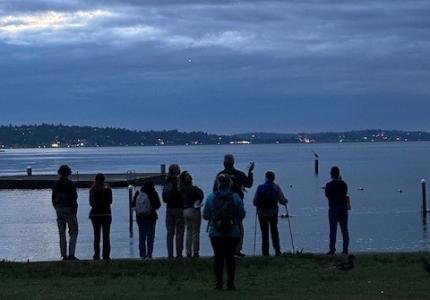 A silhouette of people standing in front of a lake at sunset. 