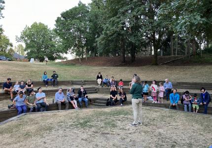 A person standing on an outdoor stage, speaking to a group of seated listeners.