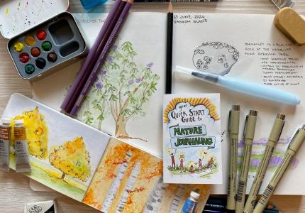 A desk with paint, markers, pens, people with a drawing of trees, and a small notebook. 