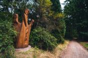 A totem-like bird carved from yellow cedar emerges from the forest along a path in a green open space.