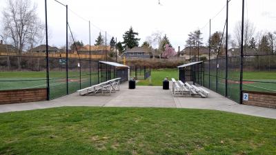 A playfield that includes a cement walkway located between two softball fields and featuring two sets of metal bleachers 
