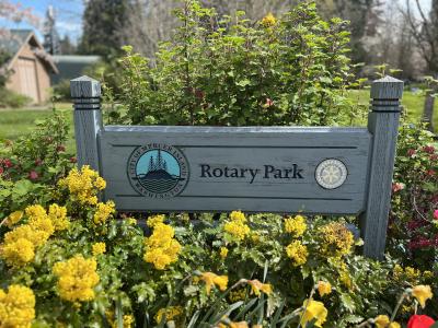 A wood sign reading Rotary Park, surrounded by yellow flowers. 