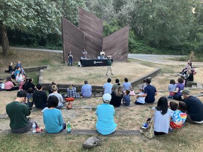 A group of people sitting in front of an amphitheater, with one speaker on stage.  