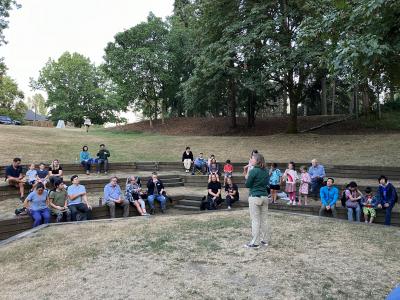 A person standing on an outdoor stage, speaking to a group of seated listeners.