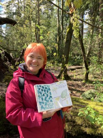 A person standing in front of a forest, holding up a notebook with drawings on it. 