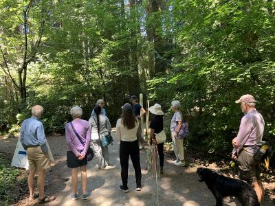 A group of people standing in a circle in the woods, looking at a speaker.
