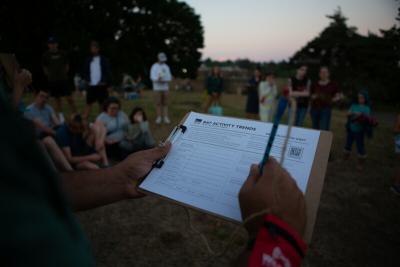 A close up of hands filling out a form on a clipboard, with people standing in the background.